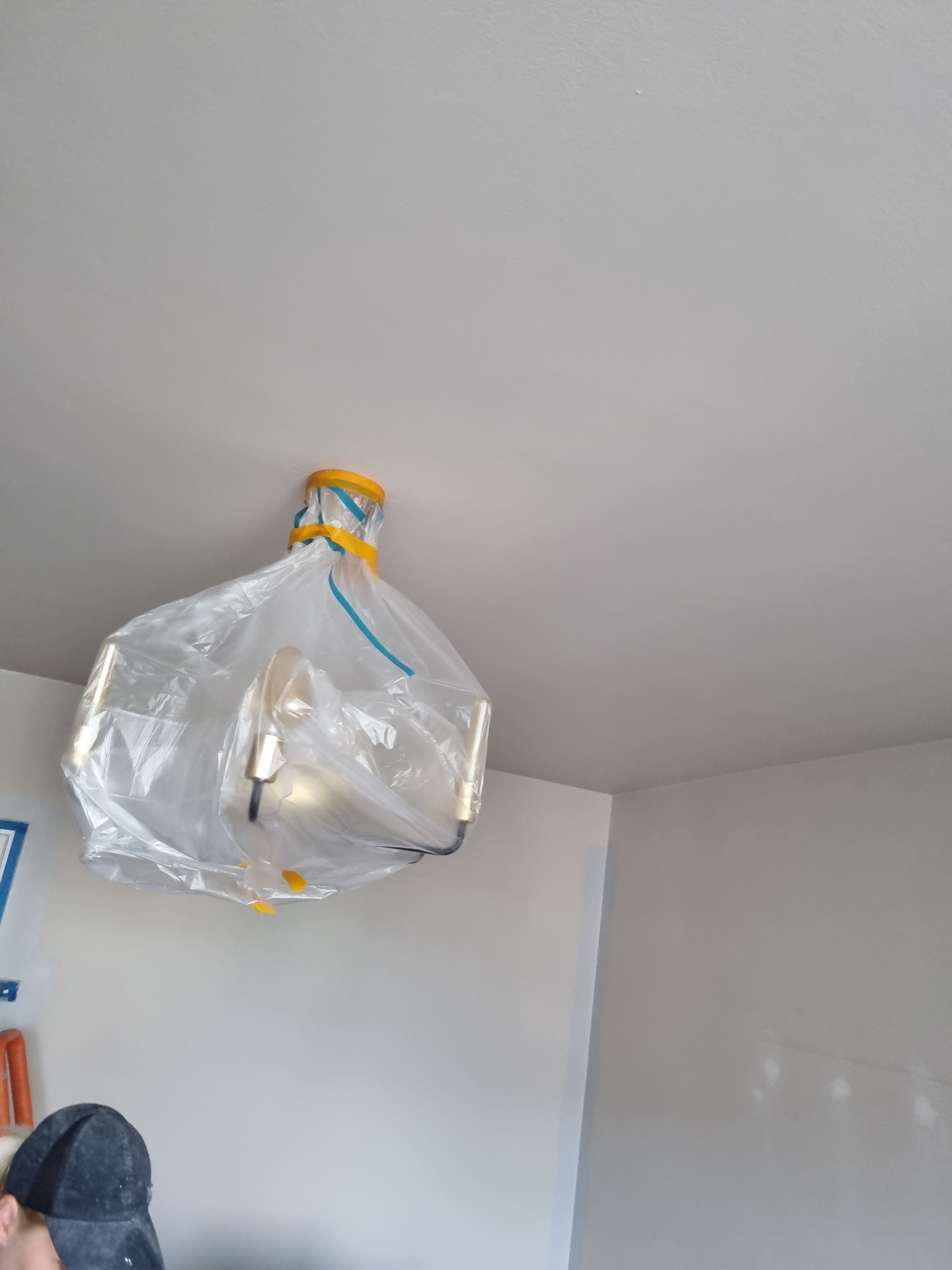 Lampa sufitowa zabezpieczona folią i taśmą malarską podczas prac malarskich, widoczny fragment ściany po nałożeniu gładzi.