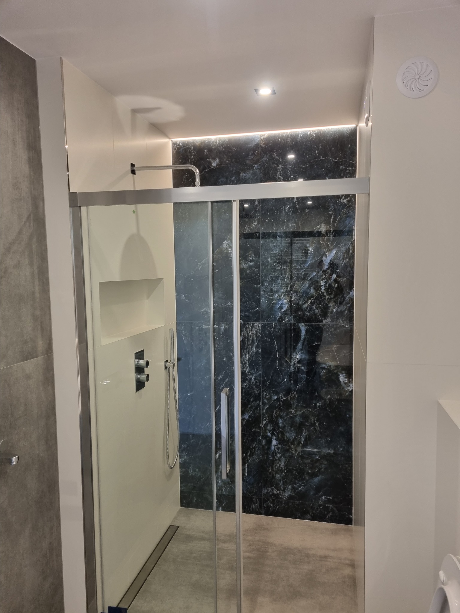 Nowoczesna kabina prysznicowa z przesuwnymi drzwiami, wykończona płytkami imitującymi czarny marmur, z wbudowaną półką i minimalistyczną armaturą.