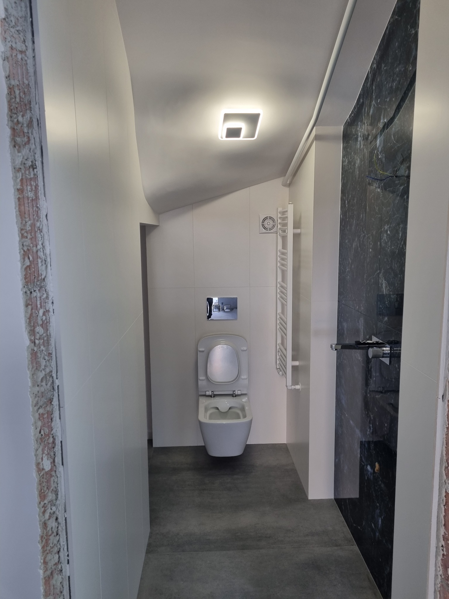 Nowoczesna łazienka z toaletą podwieszaną, grzejnikiem drabinkowym i czarną marmurową ścianą prysznicową, widok z wejścia na szare płytki podłogowe.