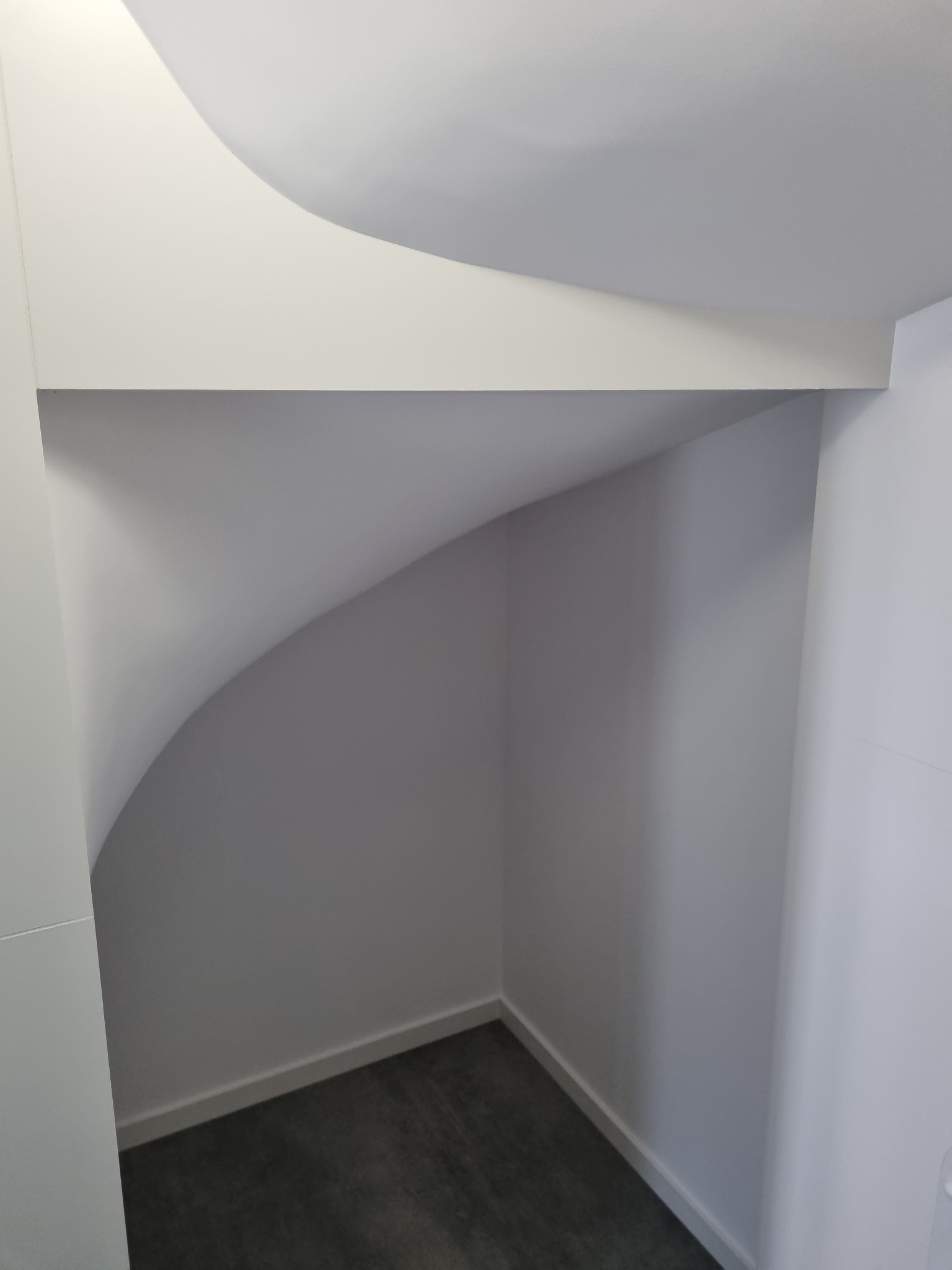 Wnęka pod schodami, wykończona w bieli, z szarą podłogą i zaokrąglonym sufitem.