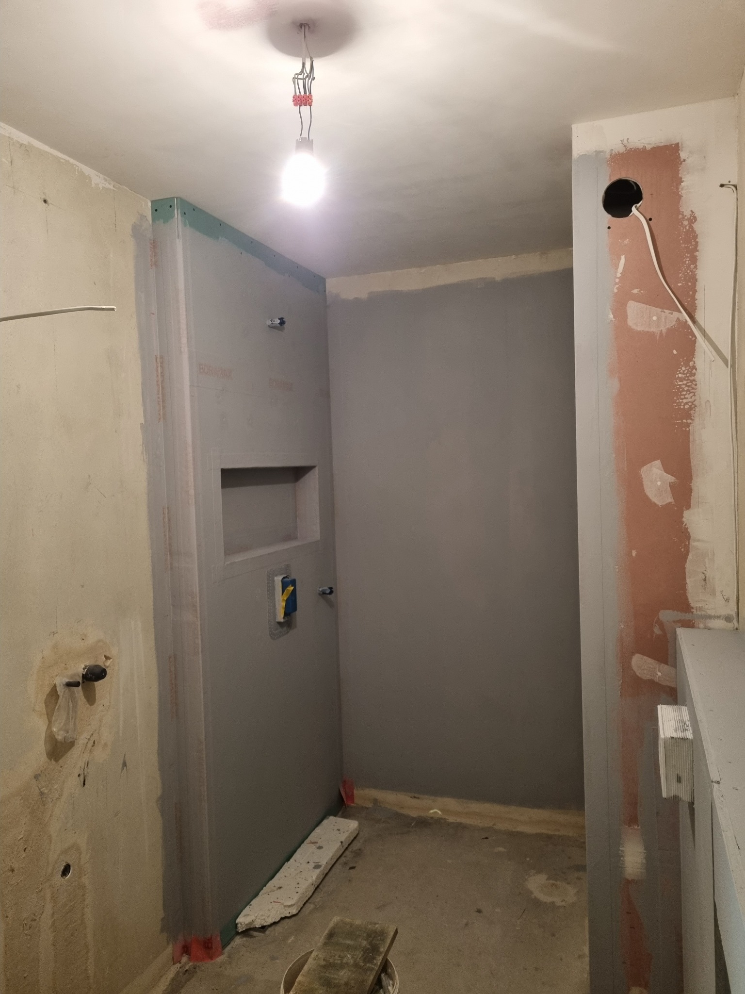 Łazienka w trakcie remontu: ściany pokryte szarą farbą, zabudowa z płyty karton-gips z wnęką, widoczne instalacje i surowy stan wykończenia.