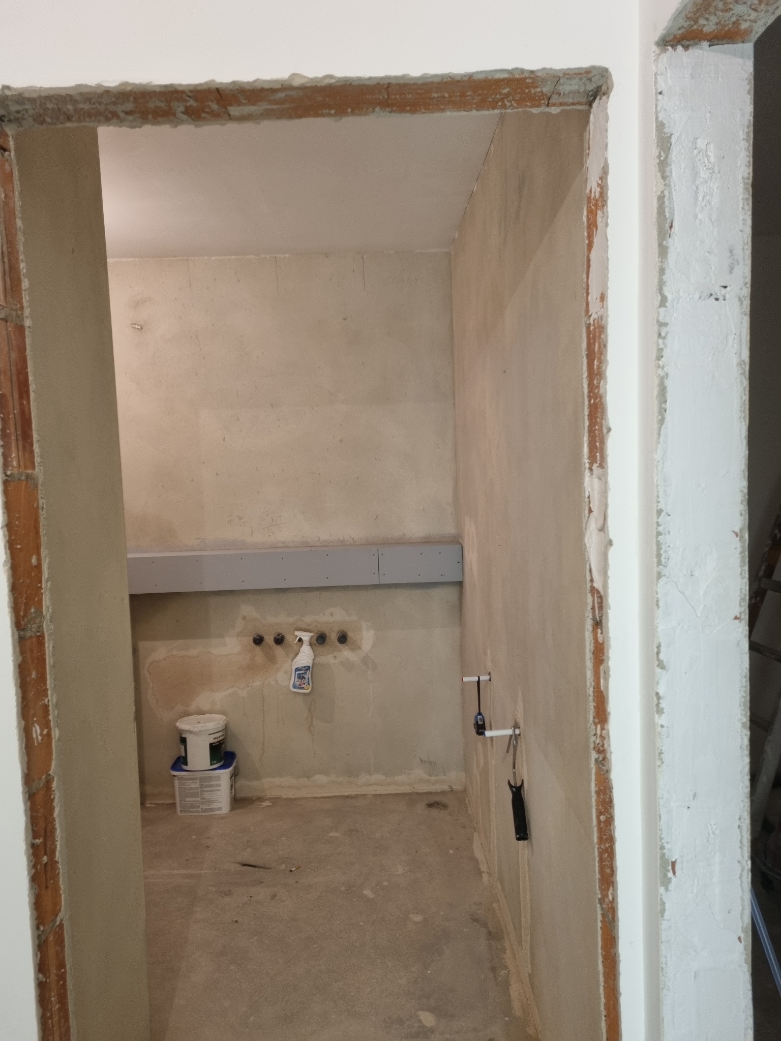 Niedokończone wnętrze: surowe ściany z widocznymi instalacjami, fragment ściany z płytą kartonowo-gipsową, wiaderko z farbą i butelka ze spryskiwaczem na podłodze, widoczne ramy drzwi w stanie...