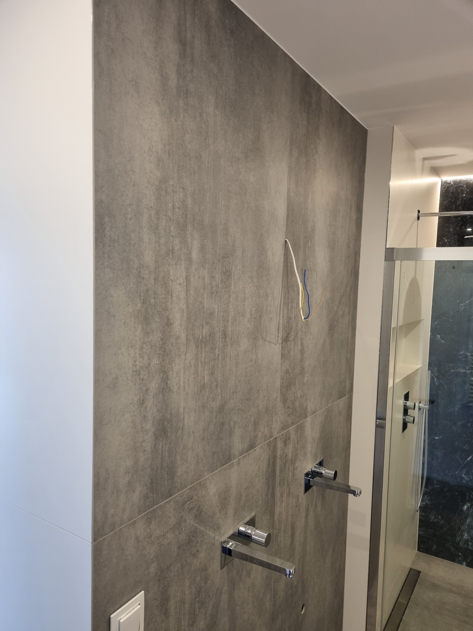 Szara ściana z płytek imitujących beton, z wystającymi przewodami elektrycznymi i zamontowaną armaturą łazienkową w nowoczesnej łazience z kabiną prysznicową.