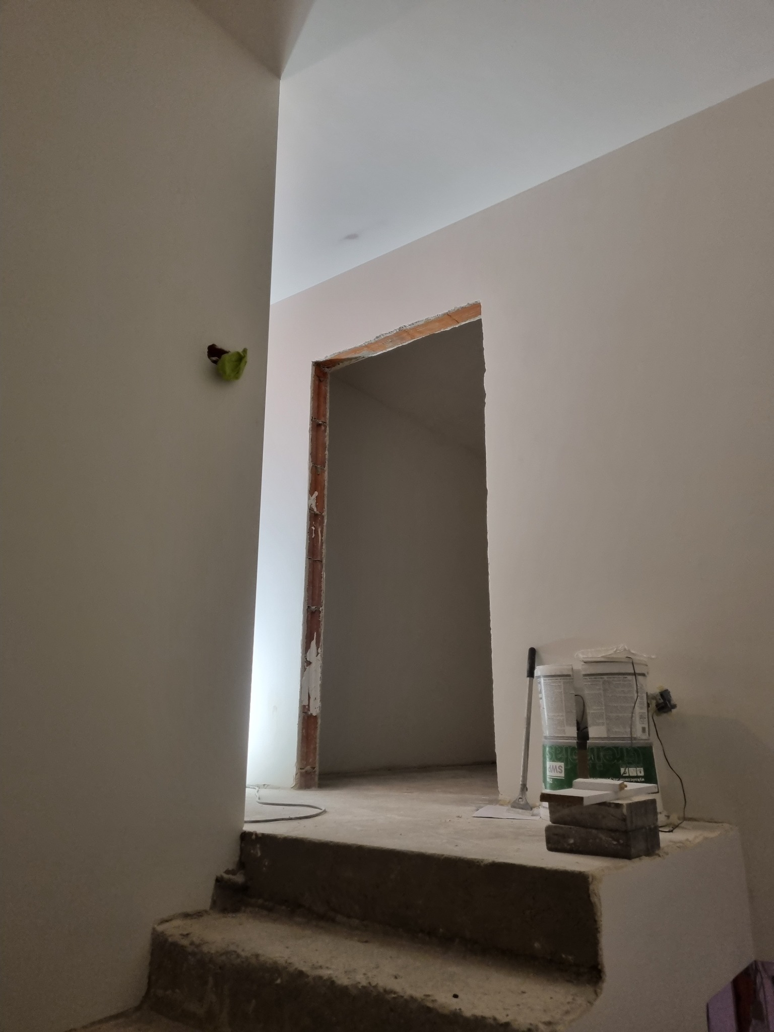 Wnętrze w trakcie prac wykończeniowych: surowe schody prowadzą do pomieszczenia z ościeżnicą bez drzwi, ściany przygotowane pod malowanie, widoczne puszki z farbą i narzędzia malarskie.