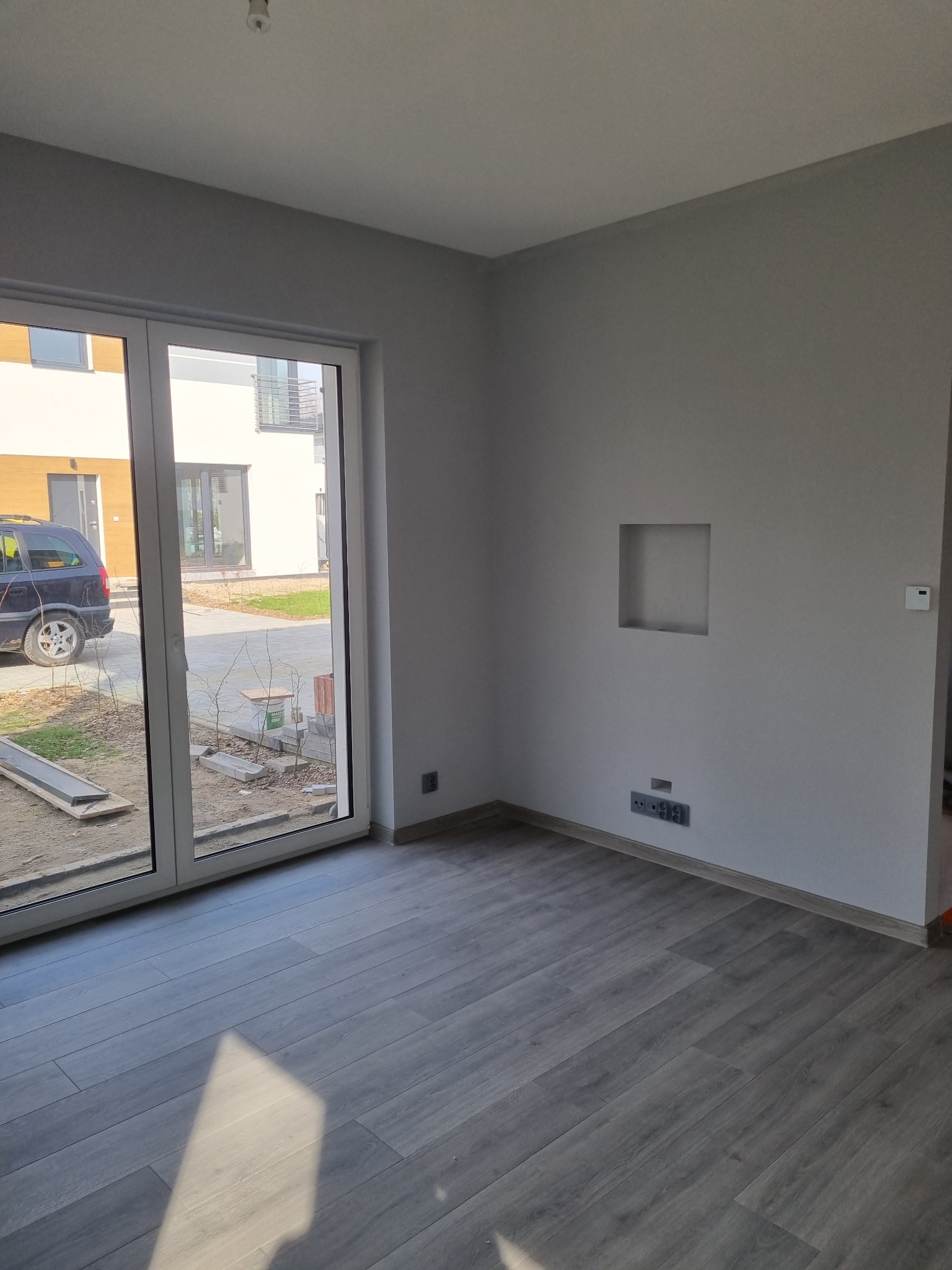Szare panele podłogowe w nowym pokoju z szarą ścianą, wnęką i widokiem na budynek przez duże okno.