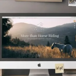 Ekran komputera iMac wyświetlający stronę internetową z motywem jeździeckim: krajobraz z koniem, logo z motywem górskim i napis 'More than Horse Riding'. Na biurku roślina w doniczce i książki.