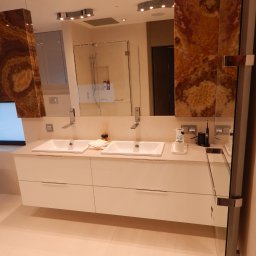 CREATIVO - Nowoczesna łazienka z dwiema umywalkami nablatowymi na białej szafce, obok wolnostojąca wanna, ściany wykończone panelami z onyksu, szklana kabina prysznicowa w tle.