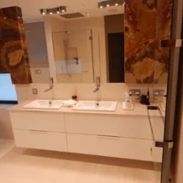 Nowoczesna łazienka z dwiema umywalkami nablatowymi na białej szafce, obok wolnostojąca wanna, ściany wykończone panelami z onyksu, szklana kabina prysznicowa w tle.