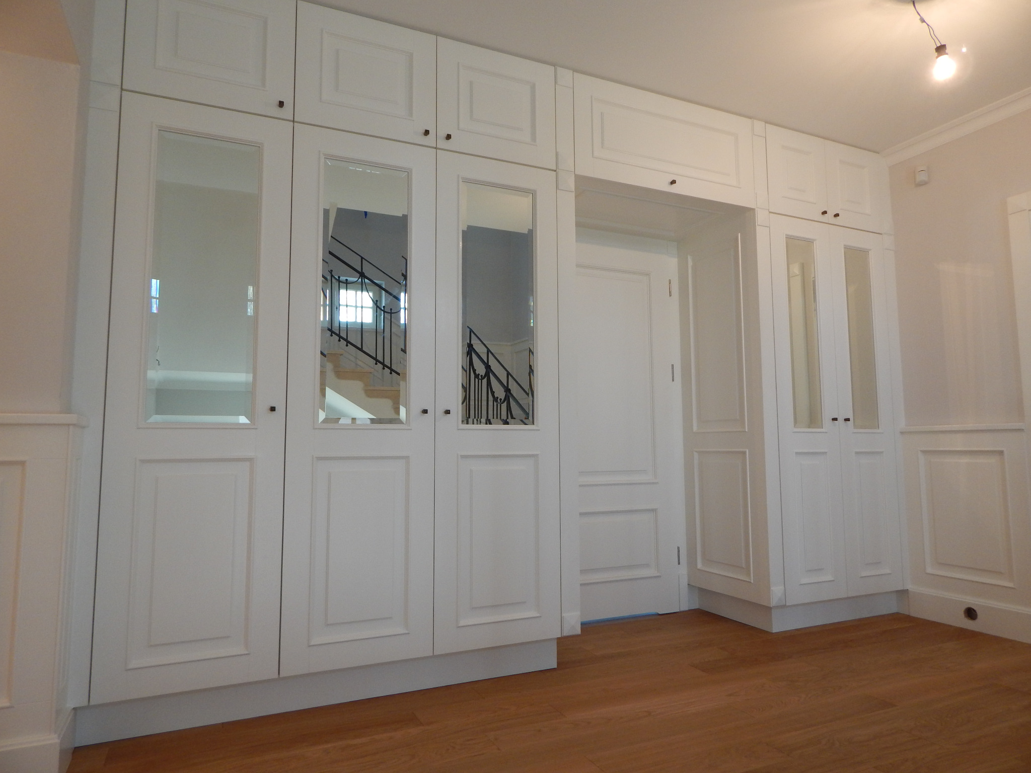 Biała szafa wnękowa z lustrami, odbijająca schody z czarną balustradą w jasnym wnętrzu, z widoczną podłogą z paneli.