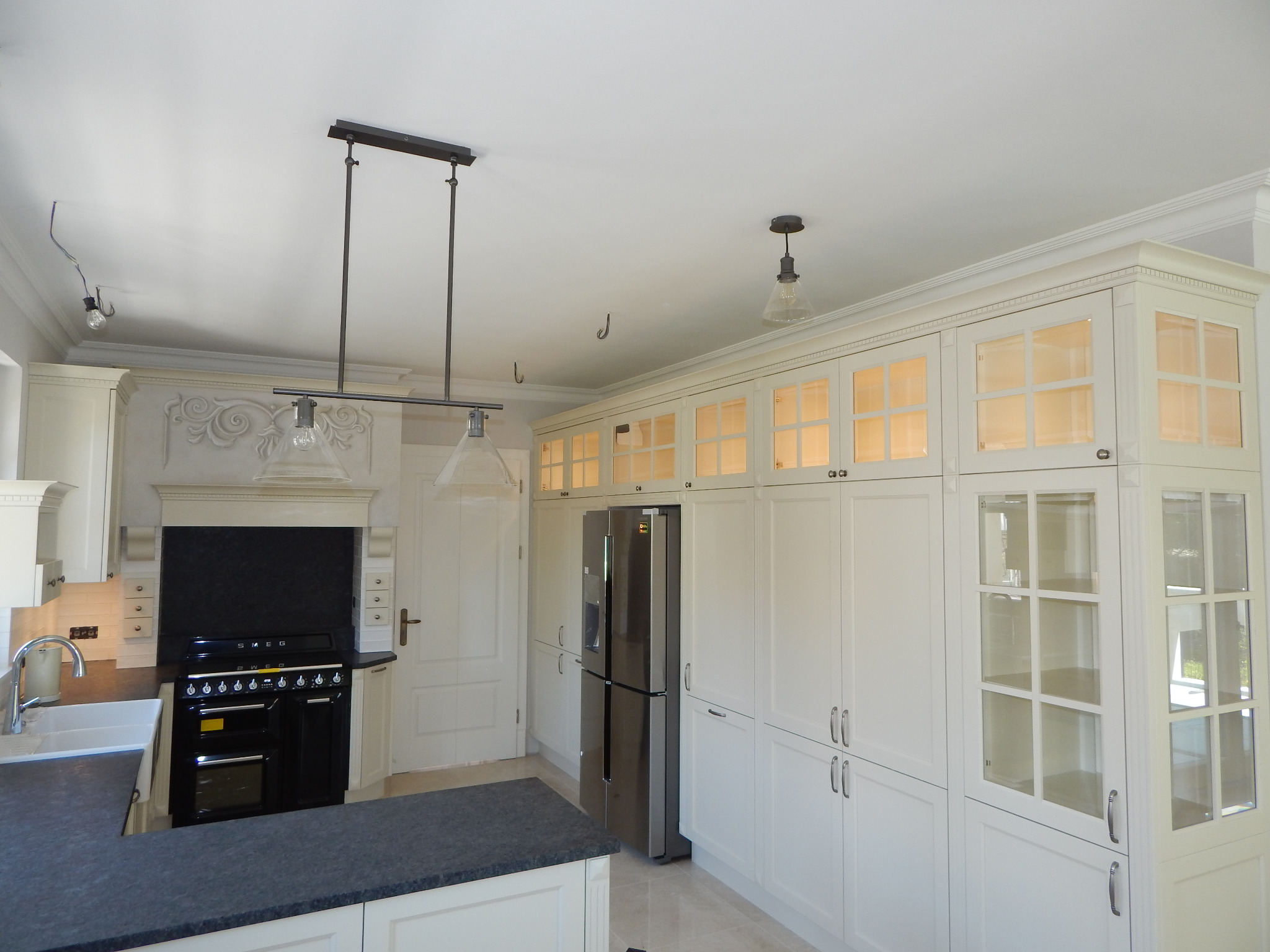 Jasna kuchnia z białymi szafkami z przeszkleniami, czarnym piekarnikiem SMEG i szarą wyspą kuchenną, uzupełniona oświetleniem w stylu industrialnym.