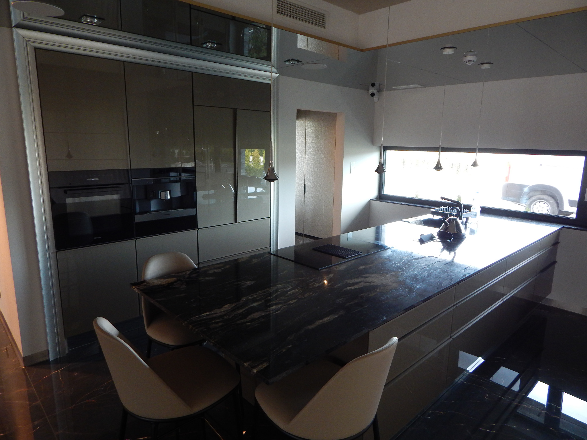 Nowoczesna kuchnia z ciemnymi, lakierowanymi frontami, wyspą z czarnym blatem z wyraźnym użyleniem oraz trzema krzesłami w odcieniach beżu. Nad wyspą zwisają minimalistyczne lampy.