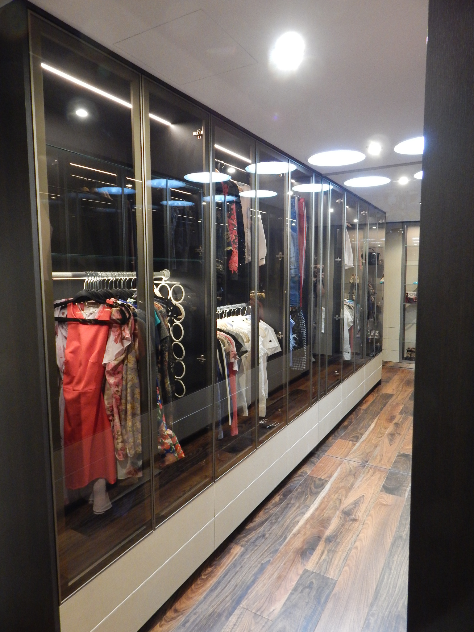 Długa, przeszklona szafa wnękowa z oświetleniem LED, eksponująca ubrania na wieszakach i półkach, z widoczną drewnianą podłogą.