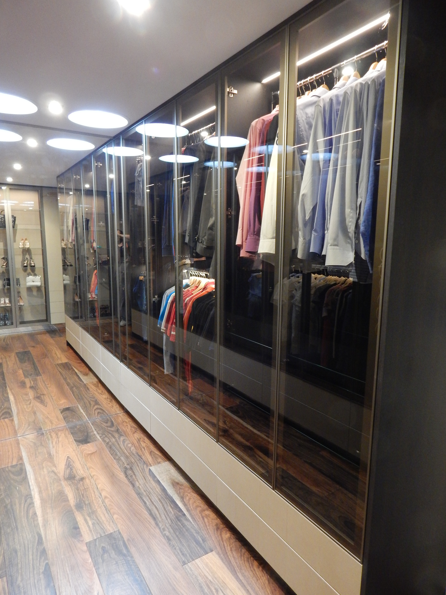 Długa szafa wnękowa z przeszklonymi drzwiami przesuwnymi, oświetleniem LED wewnątrz i drewnianą podłogą w tle.