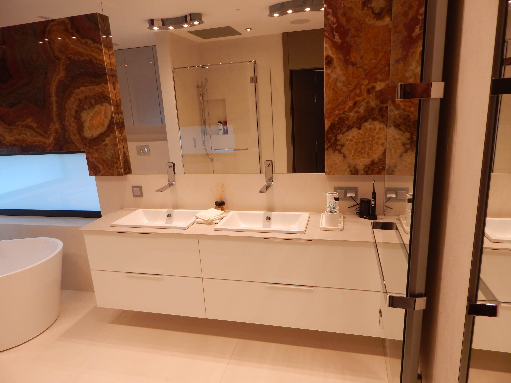 Nowoczesna łazienka z dwiema umywalkami nablatowymi na białej szafce, obok wolnostojąca wanna, ściany wykończone panelami z onyksu, szklana kabina prysznicowa w tle.