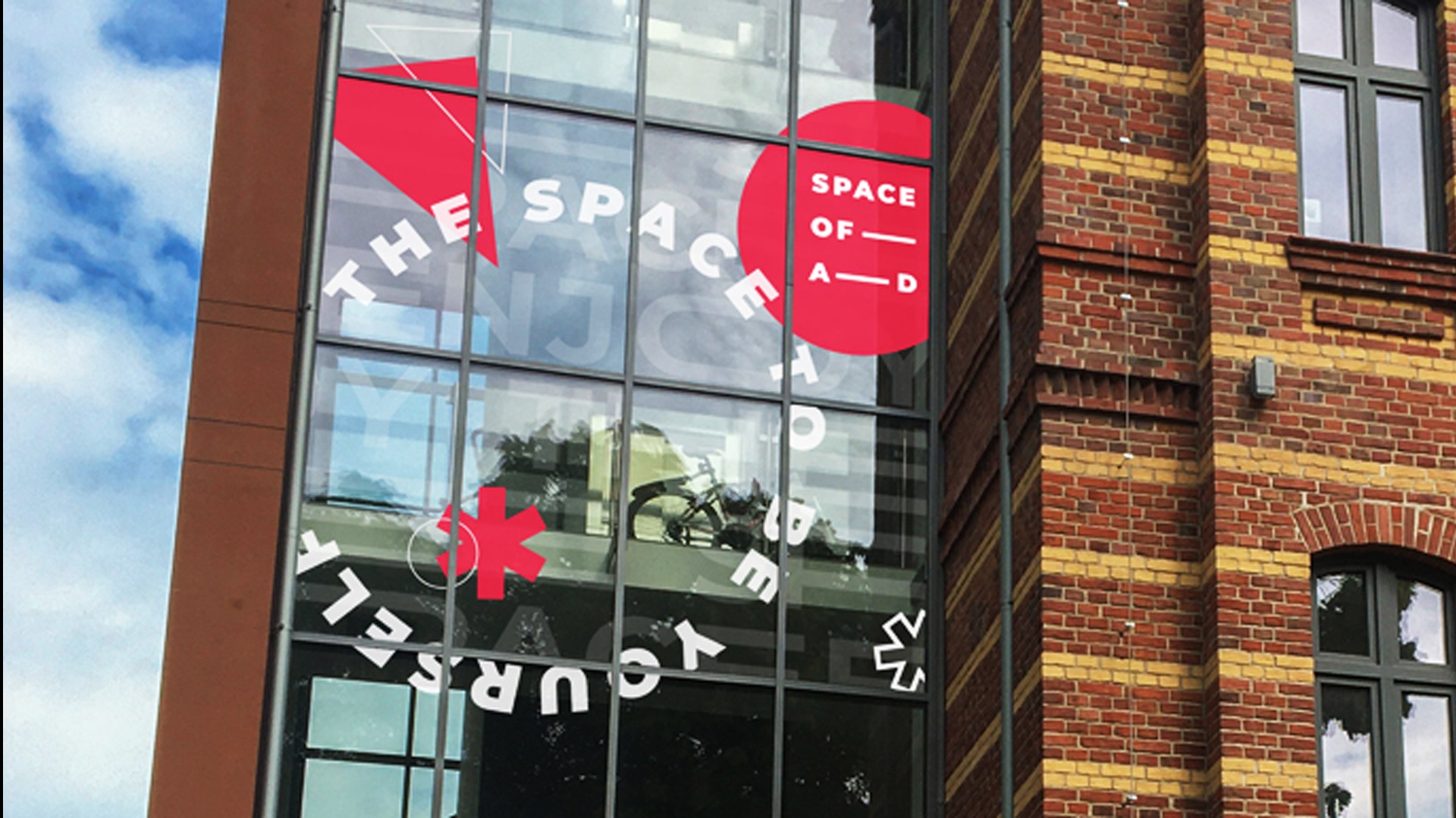 Zewnętrzna grafika na szklanej fasadzie budynku z napisem 'THE SPACE OF A-D ENJOY YOURSELF' oraz czerwonymi elementami geometrycznymi, widoczny budynek z cegły w tle.