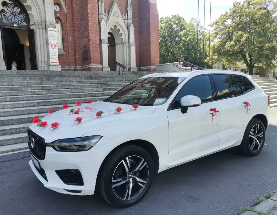 Biały SUV Volvo udekorowany czerwonymi różami i wstążkami, zaparkowany przed kościołem. Dekoracja ślubna na masce i klamkach, zielone drzewa w tle.