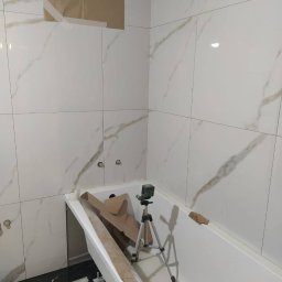 Okay Majster - Białe płytki imitujące marmur z delikatnym złotym żyłkowaniem, ułożone na ścianach łazienki w trakcie remontu, z widoczną wanną, laserowym niwelatorem na statywie, kartonowymi zabezpieczeniami...