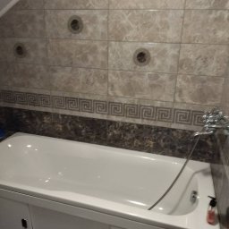 Okay Majster - Wykończona łazienka na poddaszu: biała wanna z obudową i szafkami, marmurowe płytki na ścianach z dekoracyjnym pasem z motywem greckim, trzy okrągłe otwory wentylacyjne w górnej części ściany.