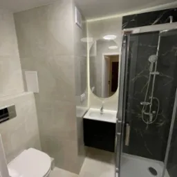 Wyremontowana łazienka z kabiną prysznicową z czarnym marmurem, wiszącą szafką z umywalką i owalnym lustrem podświetlonym LED, wykończona płytkami w odcieniach szarości.