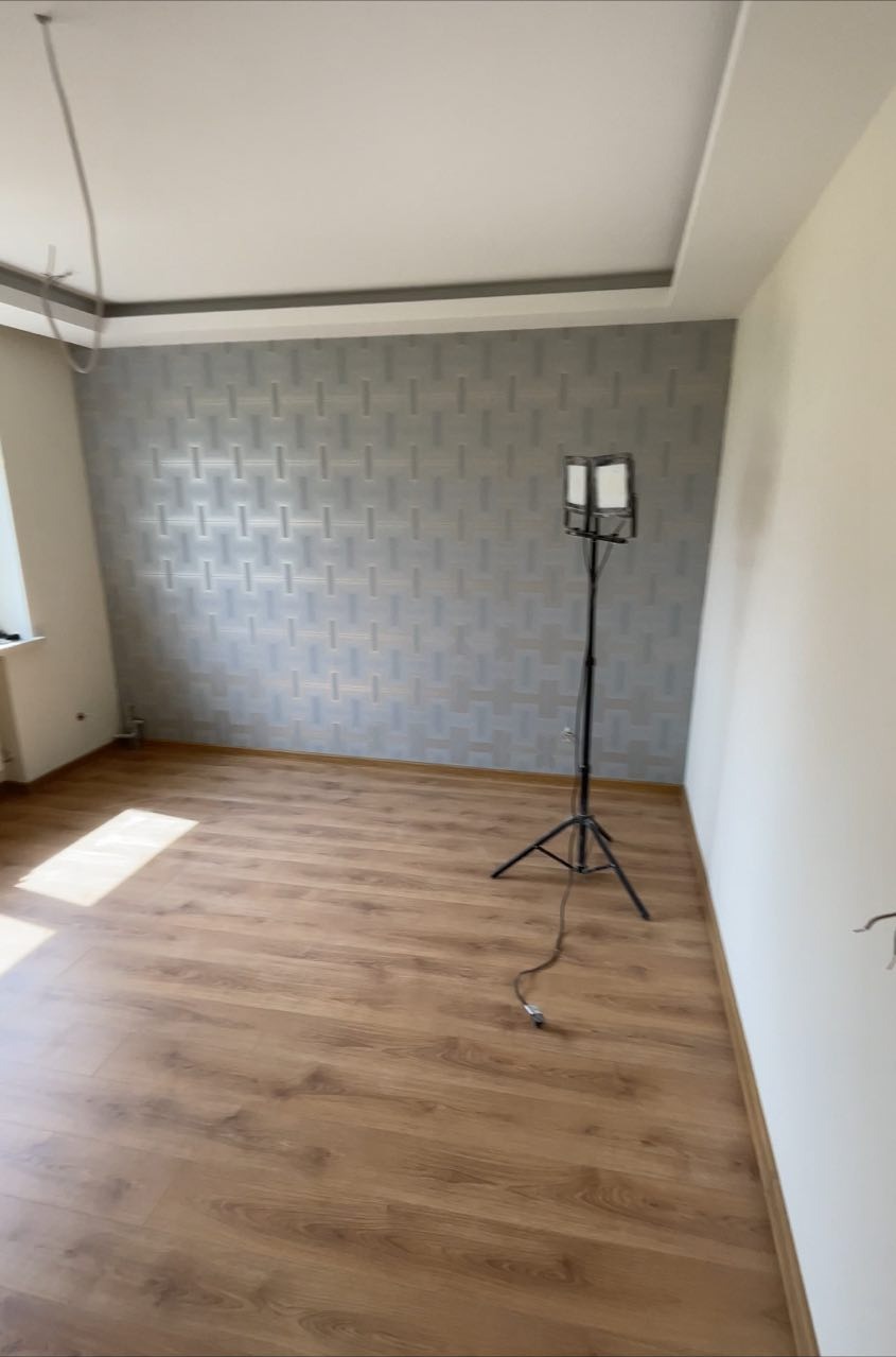 Pokój po remoncie z nową tapetą w odcieniach szarości i brązu, panele podłogowe w ciepłym odcieniu drewna, widoczna lampa na statywie.