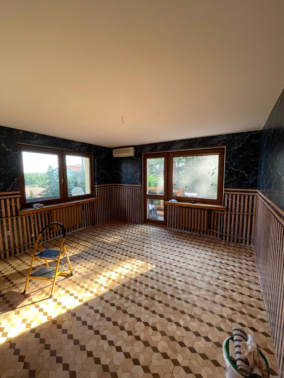 Pokój po remoncie z wzorzystą podłogą, ciemnymi ścianami z drewnianą boazerią i drabinką malarską w świetle słonecznym.