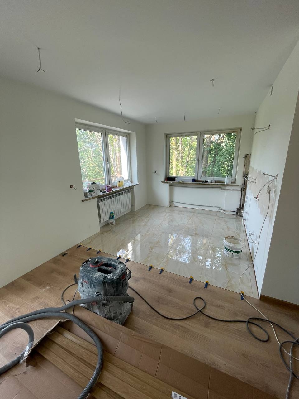 Fragment pokoju w trakcie układania paneli podłogowych, widoczny odkurzacz przemysłowy, płytki ceramiczne na podłodze w tle, niebieskie kliny dystansowe pomiędzy panelami.