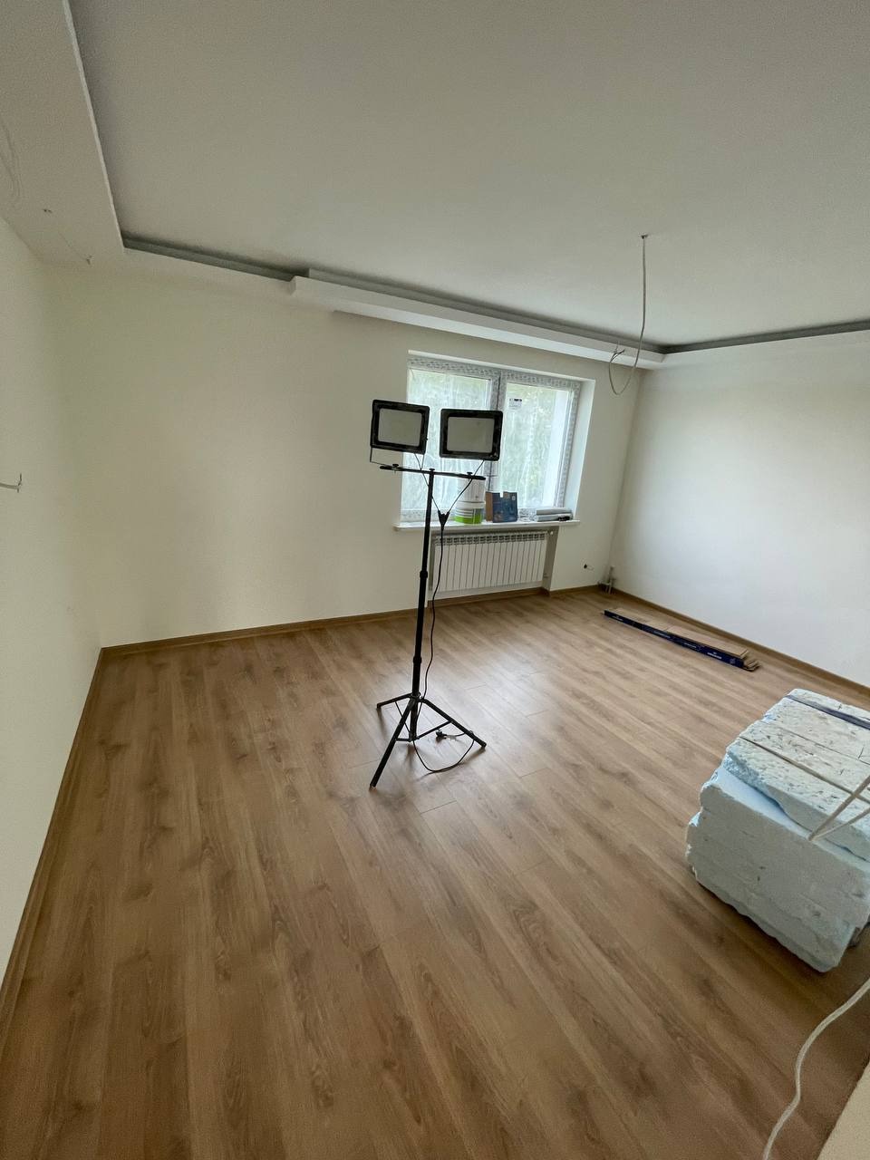 Pokój po ułożeniu paneli podłogowych, widoczne białe ściany, okno z grzejnikiem, lampa na statywie oraz materiały izolacyjne w rogu, sufit z listwami.