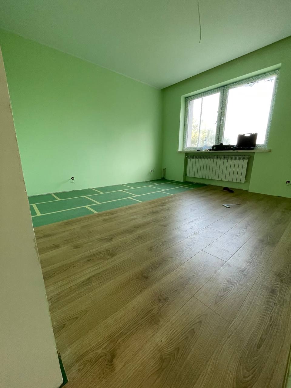 Pokój z zielonymi ścianami w trakcie układania paneli podłogowych; widoczna część ułożonej podłogi oraz zielona folia pod panele, grzejnik pod oknem.