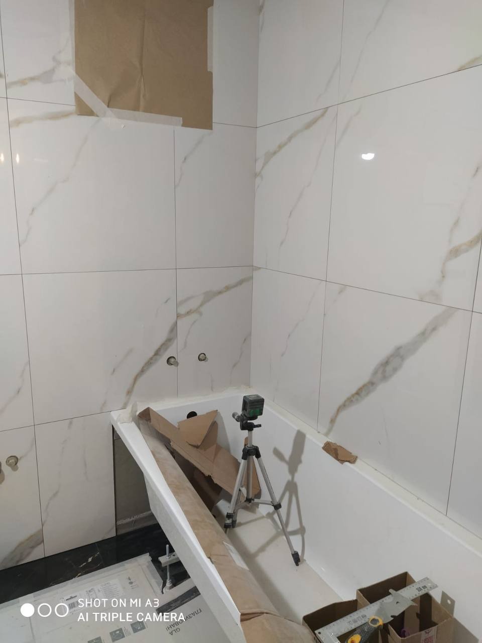 Białe płytki imitujące marmur z delikatnym złotym żyłkowaniem, ułożone na ścianach łazienki w trakcie remontu, z widoczną wanną, laserowym niwelatorem na statywie, kartonowymi zabezpieczeniami...