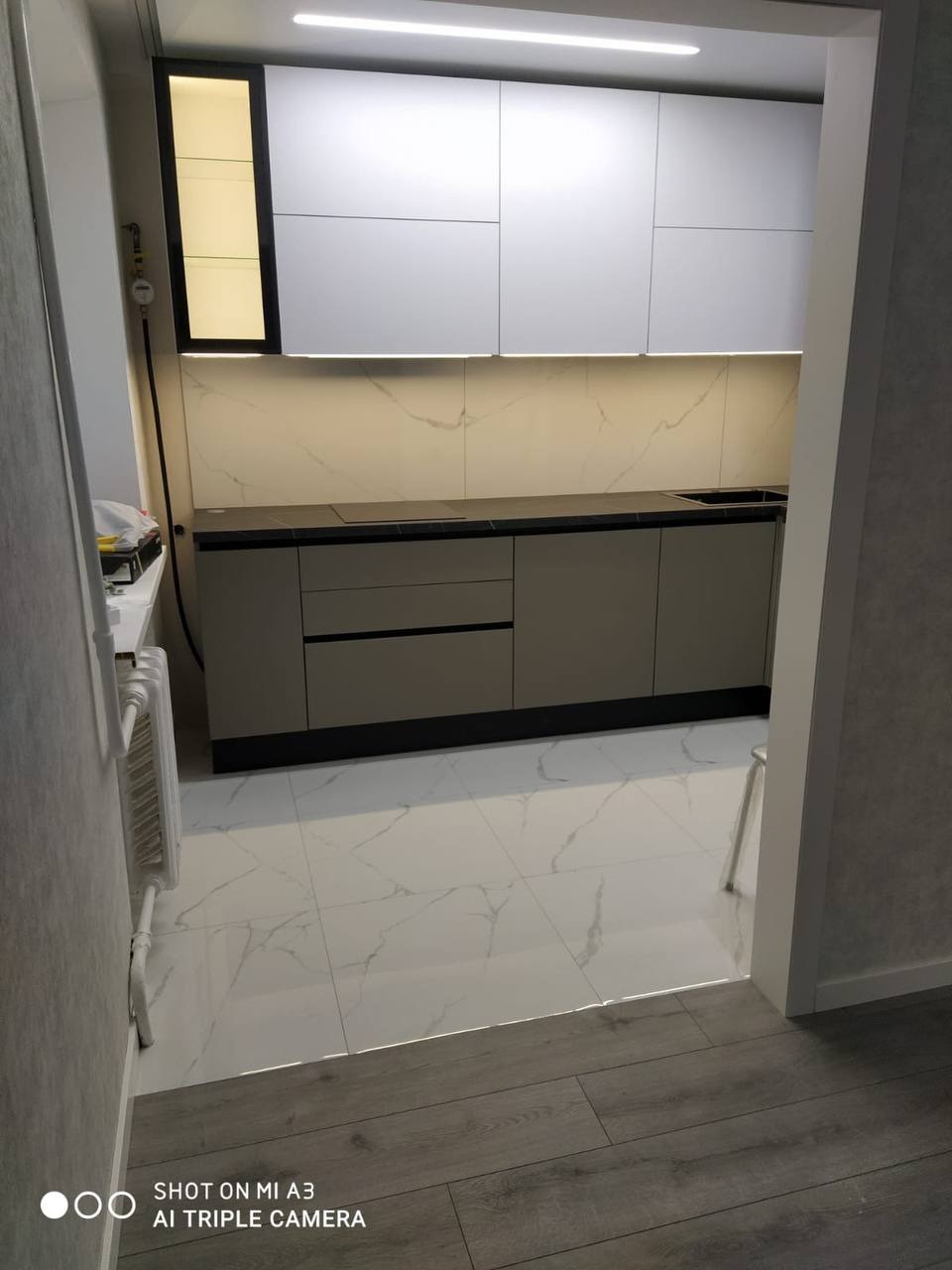 Nowoczesna kuchnia w odcieniach szarości i bieli, z marmurowym wzorem na ścianie, oświetleniem LED pod szafkami i białymi płytkami podłogowymi z szarymi żyłkami, widziana z perspektywy wejścia.