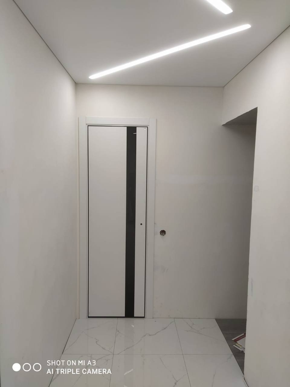 Nowoczesne wnętrze: białe ściany, marmurowe płytki podłogowe i białe drzwi z czarnym pasem, oświetlenie LED na suficie.