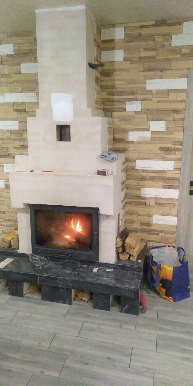 Wnętrze z kominkiem w trakcie wykończenia, widoczne płyty imitujące kamień na ścianie, szpachlowany kominek z paleniskiem, podłoga wyłożona płytkami imitującymi drewno, oraz torba budowlana w rogu