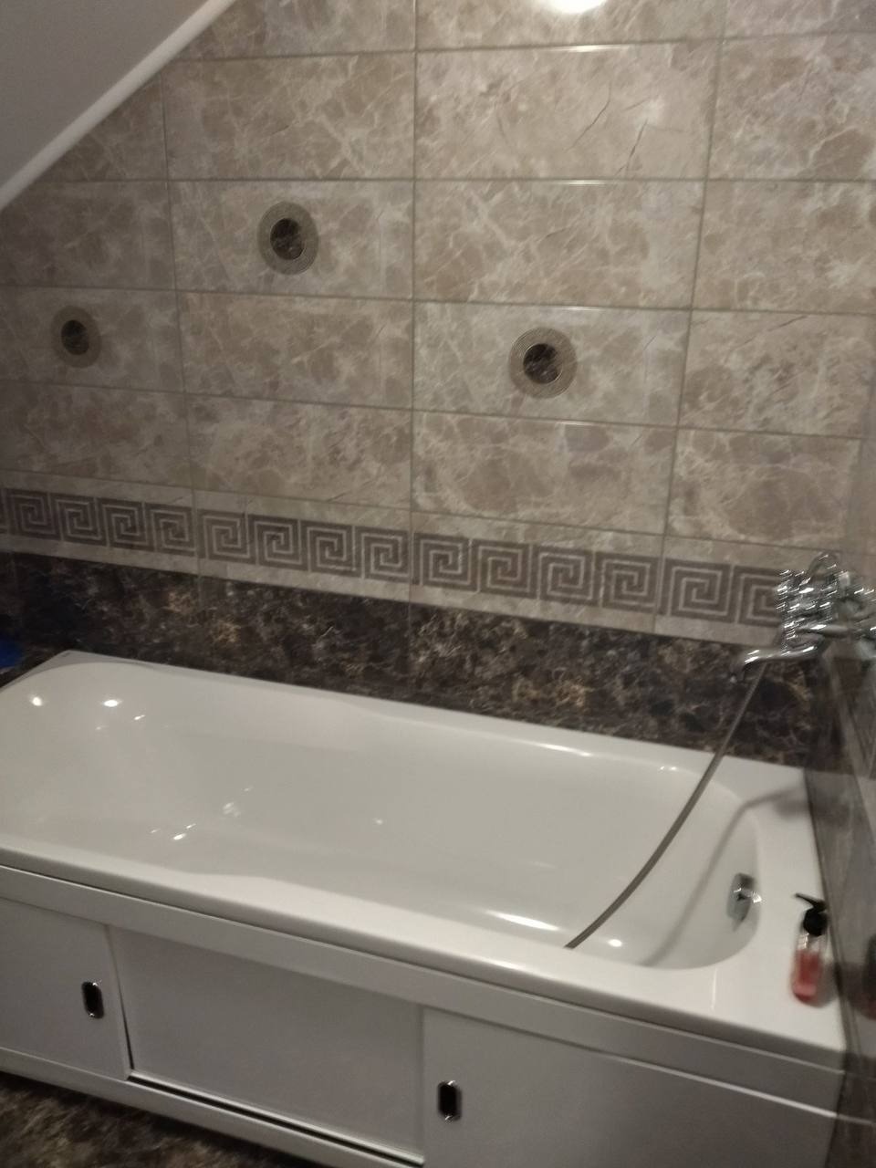 Wykończona łazienka na poddaszu: biała wanna z obudową i szafkami, marmurowe płytki na ścianach z dekoracyjnym pasem z motywem greckim, trzy okrągłe otwory wentylacyjne w górnej części ściany.