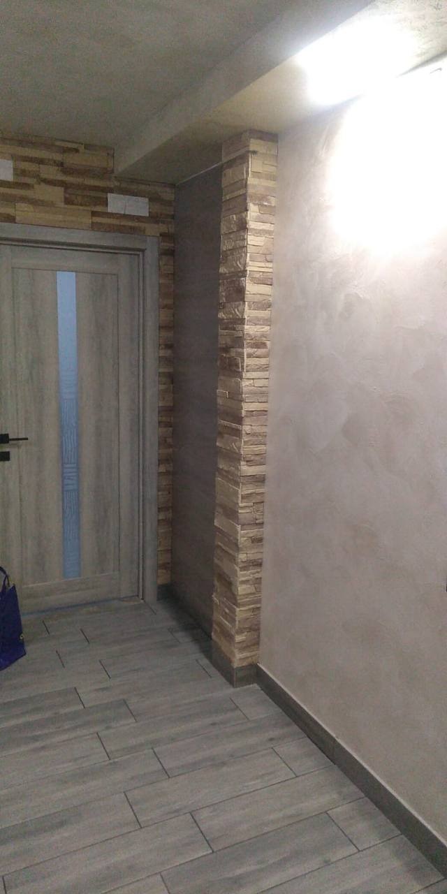 Wykończony korytarz z szarymi panelami podłogowymi imitującymi drewno, dekoracyjną ścianą z kamienia elewacyjnego na filarze oraz ścianą wykończoną tynkiem dekoracyjnym w odcieniach beżu i szarości...