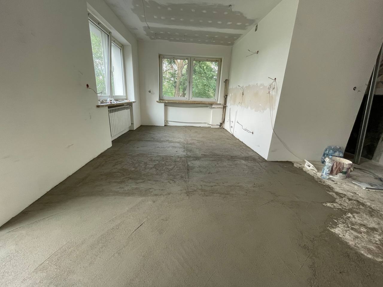 Wnętrze pokoju w trakcie wylewki podłogi, widoczne okna, grzejniki, ściany z częściowo położoną gładzią, wystające kable elektryczne i narzędzia budowlane.