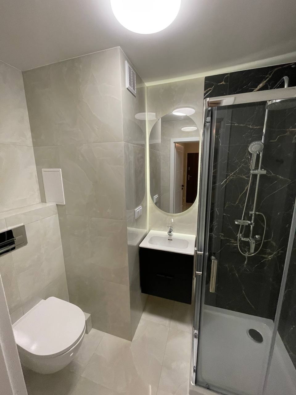 Wyremontowana łazienka z kabiną prysznicową z czarnym marmurem, wiszącą szafką z umywalką i owalnym lustrem podświetlonym LED, wykończona płytkami w odcieniach szarości.
