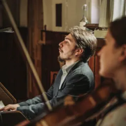 Młody mężczyzna w szarym garniturze gra na organach w kościele, obok niego skrzypaczka podczas ceremonii ślubnej.