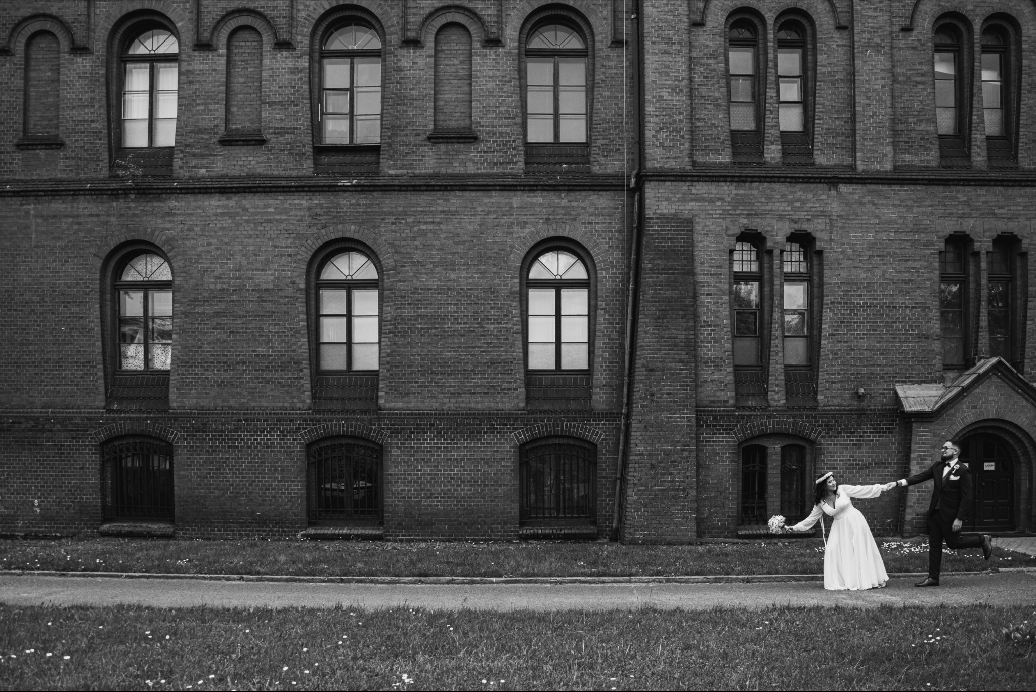 Czarno-białe zdjęcie pary młodej trzymającej się za ręce, biegnącej przed ceglanym budynkiem z łukowymi oknami i trawnikiem w tle.