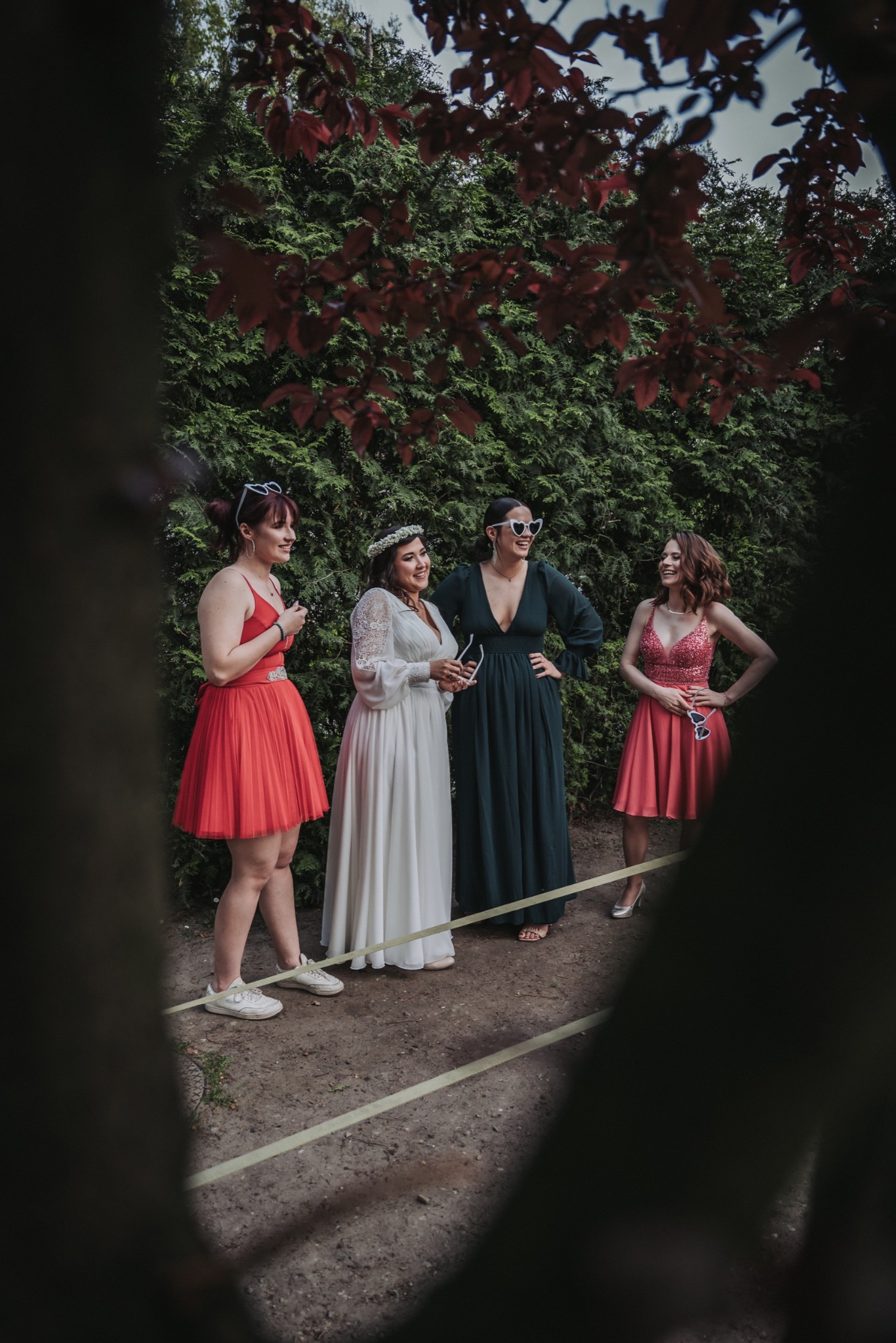 Panna młoda w koronkowej sukni i druchny w czerwonych i zielonej sukni, wszystkie w okularach w kształcie serc, stoją na zewnątrz na tle zielonego żywopłotu.