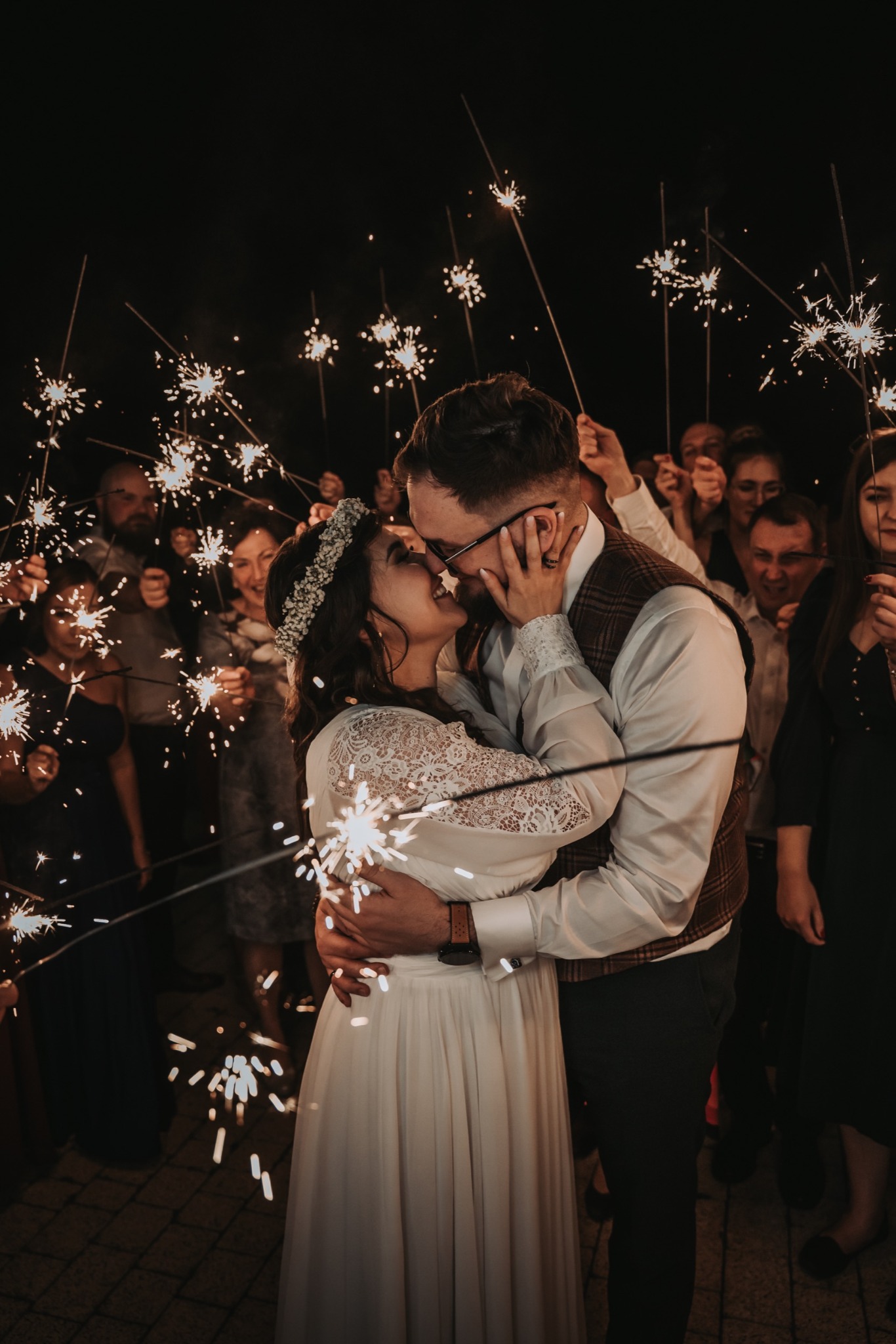 Para młoda całuje się otoczona przez gości weselnych trzymających płonące zimne ognie, ujęcie w ciemności, delikatne oświetlenie podkreśla radość chwili.