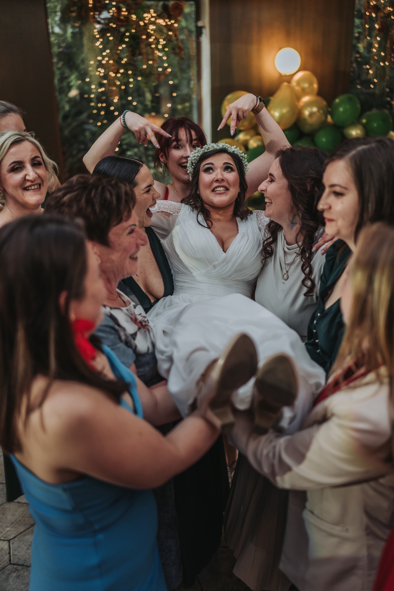 Uśmiechnięta panna młoda w białej sukni i wianku unoszona przez rozbawione druhny na tle dekoracji ze światełek i balonów.