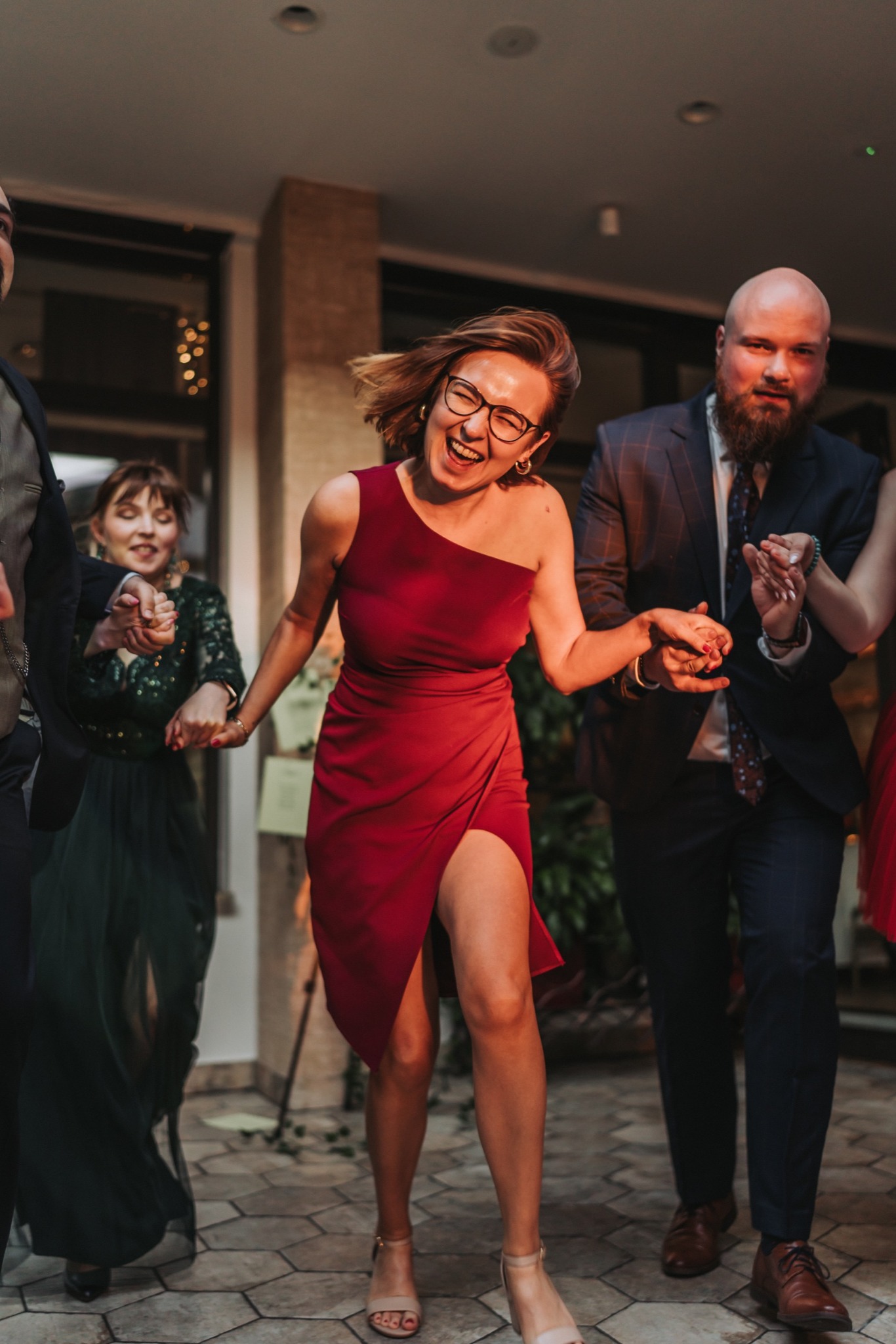 Uczestnicy wesela trzymający się za ręce, kobieta w czerwonej sukience z rozcięciem śmieje się, mężczyzna w granatowym garniturze z brodą patrzy w obiektyw, taniec na parkiecie.