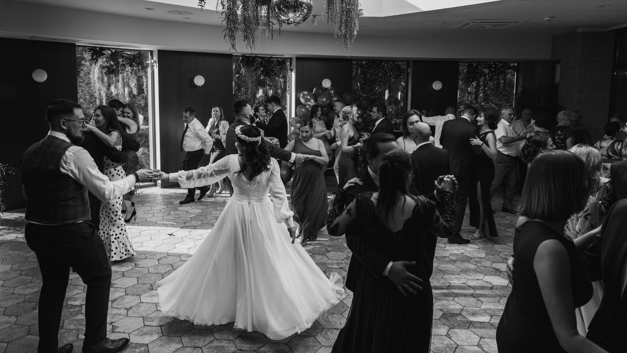 Czarno-białe zdjęcie sali balowej z tańczącymi gośćmi weselnymi; panna młoda w sukni z długimi rękawami wiruje na parkiecie z geometrycznym wzorem.