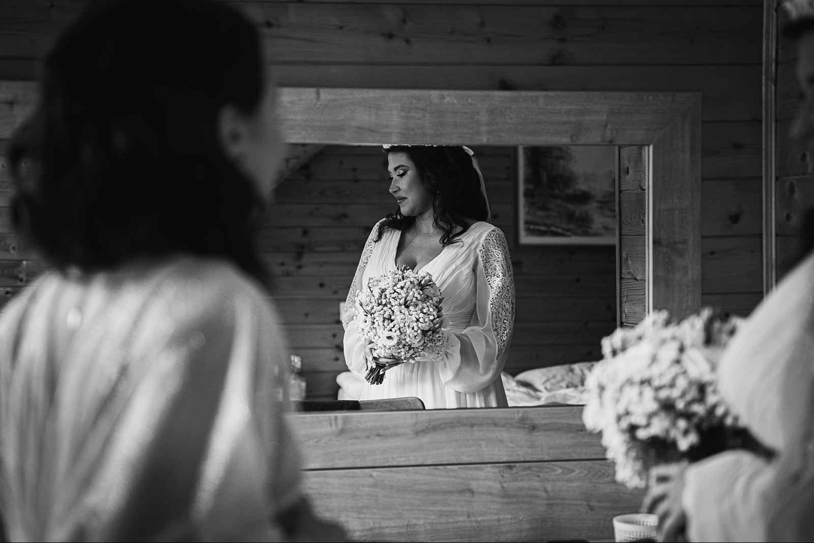 Czarno-białe zdjęcie panny młodej w lustrze, trzymającej bukiet, z drewnianym tłem i fragmentami innych osób w kadrze.