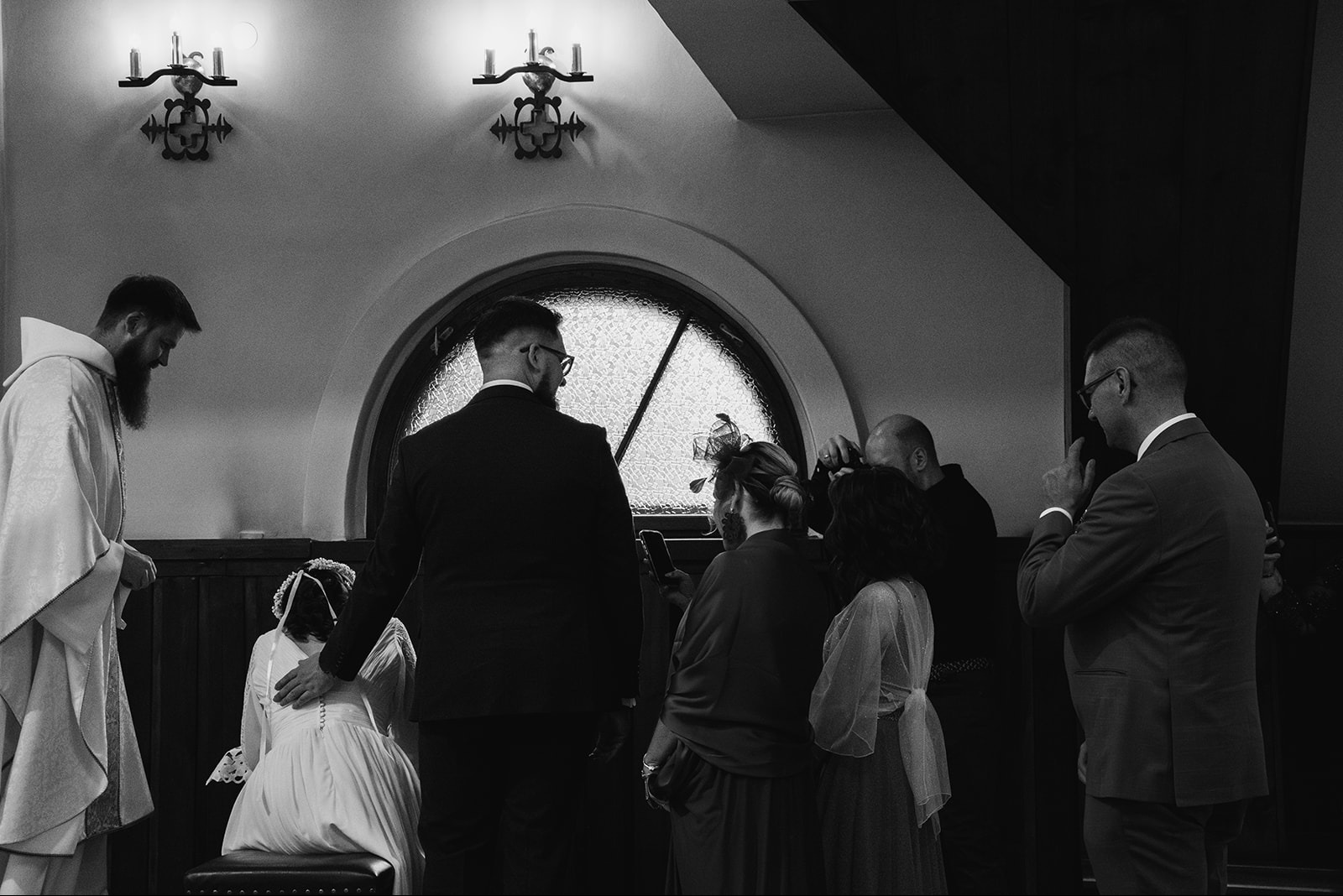 Czarno-białe zdjęcie z wnętrza kościoła, ukazujące klęczącą pannę młodą w białej sukni, księdza z brodą, mężczyznę w garniturze kładącego dłoń na jej plecach, oraz grupę gości obserwujących scenę...