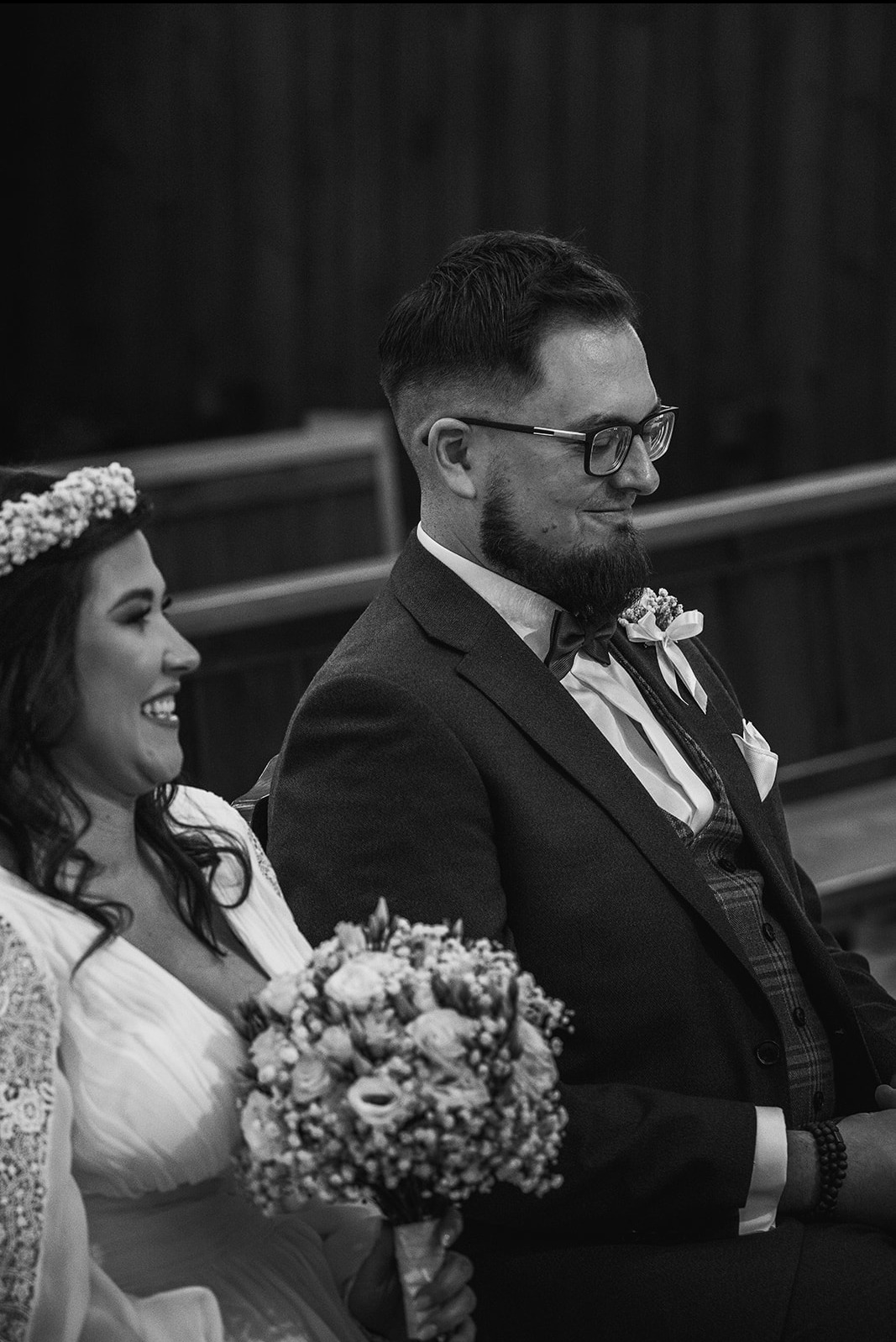 Czarno-białe ujęcie uśmiechniętej panny młodej w wianku i eleganckiego pana młodego w okularach, siedzących obok siebie podczas ceremonii.