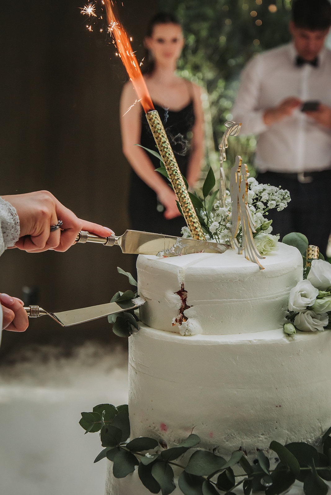 Dwie osoby kroją tort weselny nożami o zdobionych rękojeściach, z fontanną iskier umieszczoną na szczycie, w tle para gości.