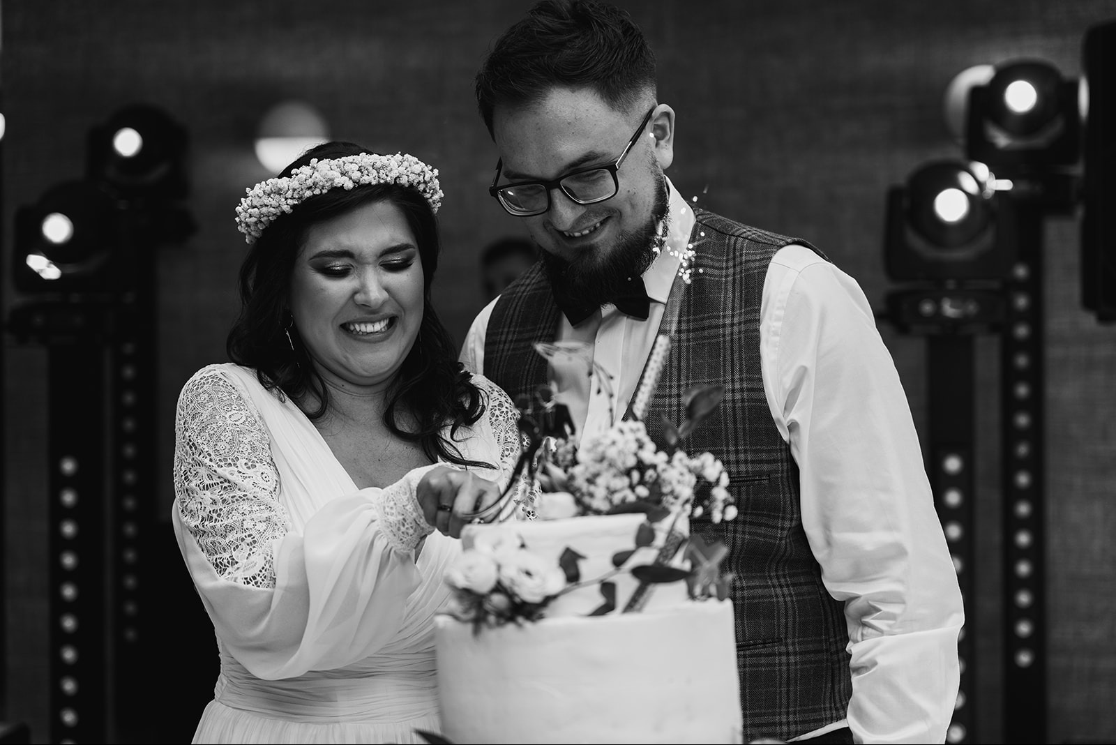 Czarno-białe zdjęcie uśmiechniętej pary młodej krojącej tort weselny, panna młoda w koronie z kwiatów i sukni z koronkowymi rękawami, pan młody w okularach i kraciastej kamizelce, w tle oświetlenie...