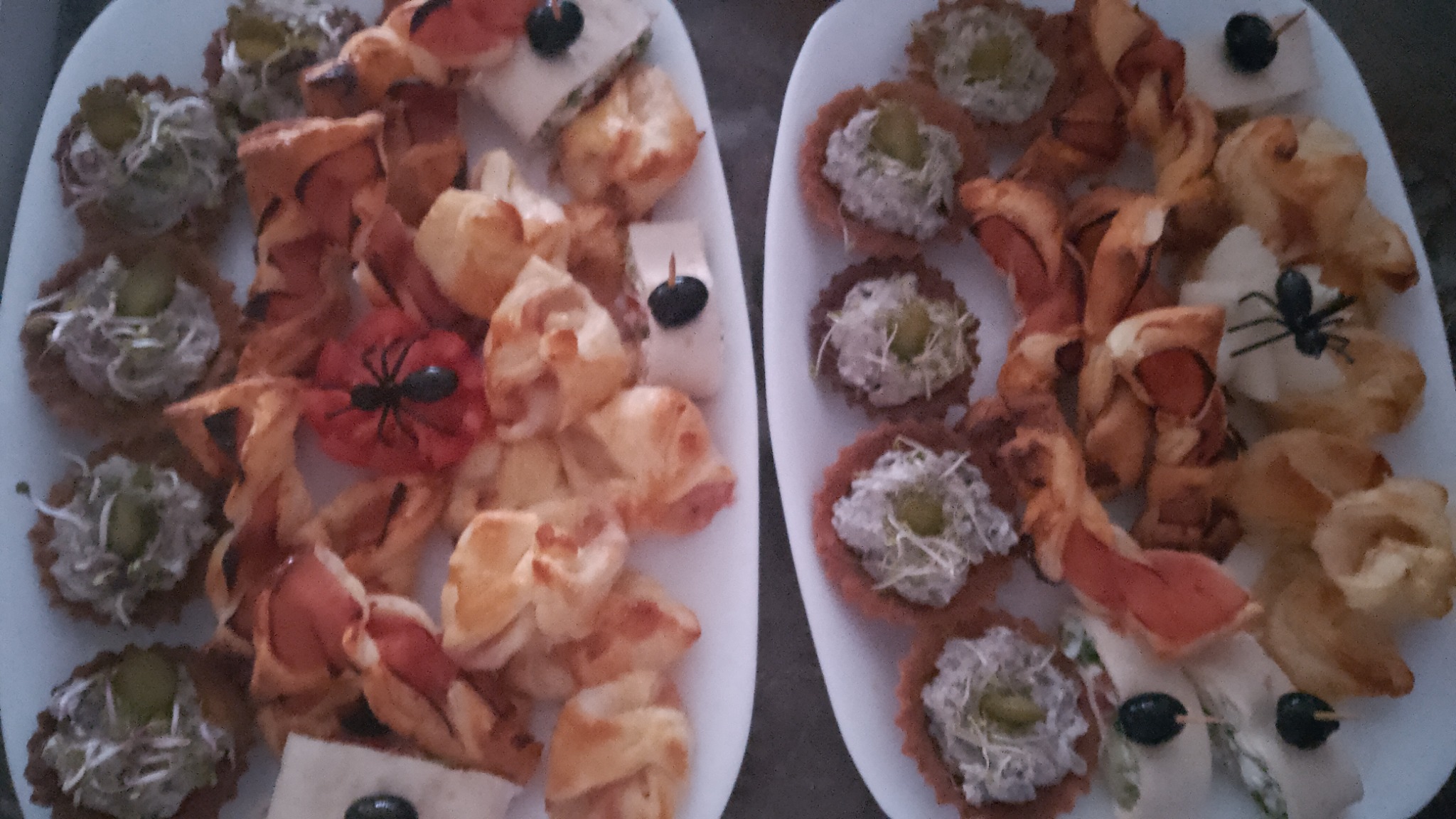 Dwa białe talerze z różnorodnymi przekąskami: tartaletki z pastą i ogórkiem, roladki z ciasta francuskiego z szynką, kanapeczki z serem i oliwką, niektóre udekorowane plastikowym pająkiem.