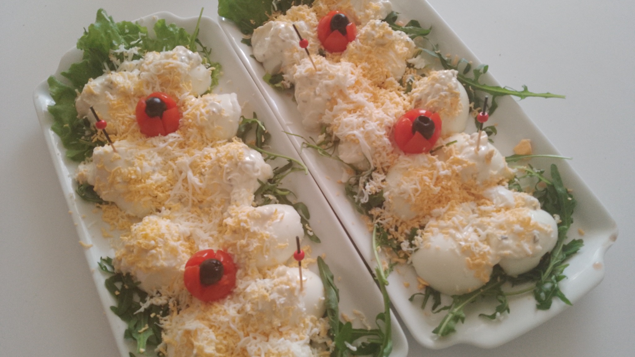 Dwie prostokątne patery z jajkami w majonezie posypanymi startym żółtkiem, udekorowane sałatą, rukolą i pomidorkami koktajlowymi z oliwką, umieszczonymi na wykałaczkach.